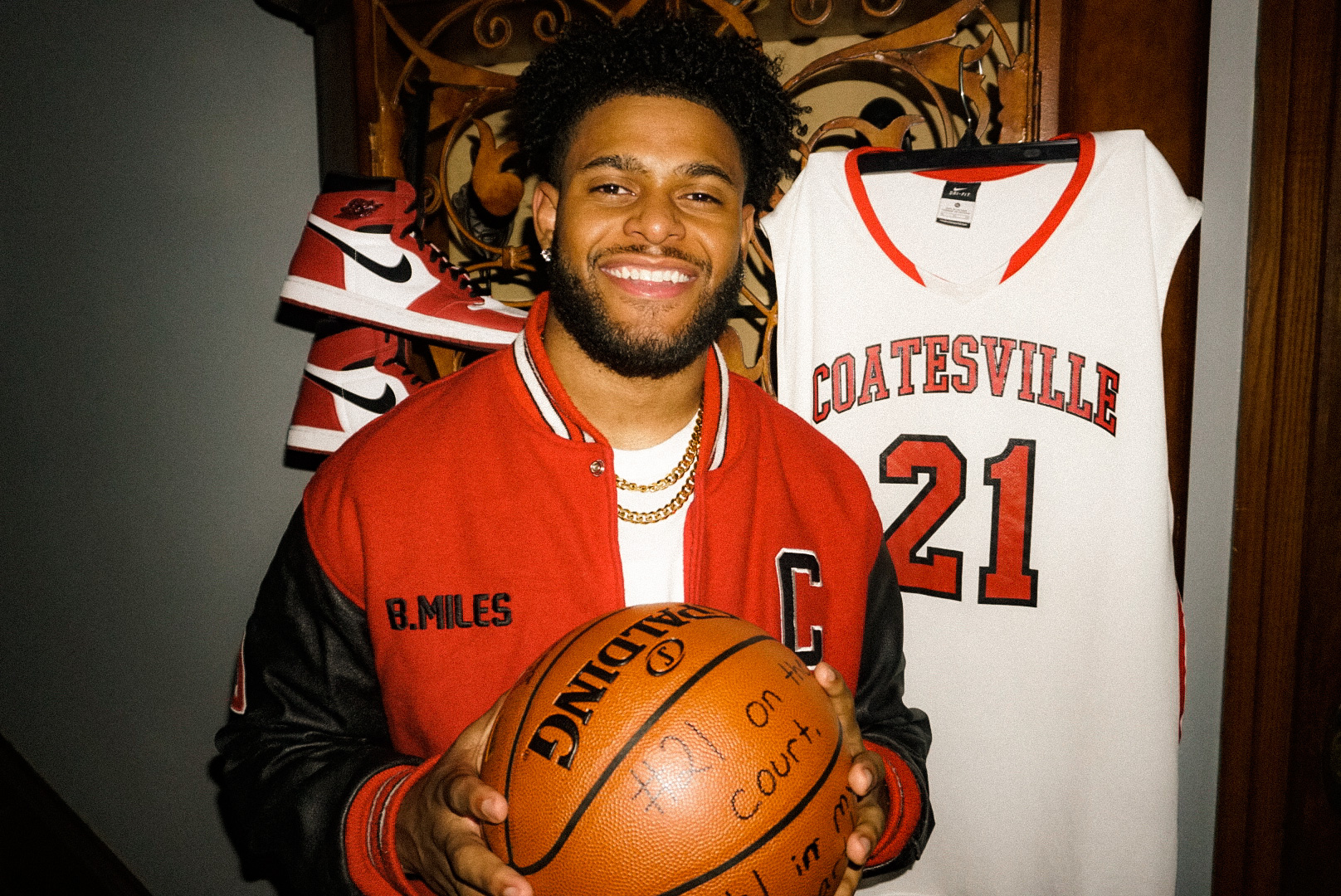 Brandon Miles holding a basketball in his red and black Coatesville letterman jacket, beside his number 21 jersey